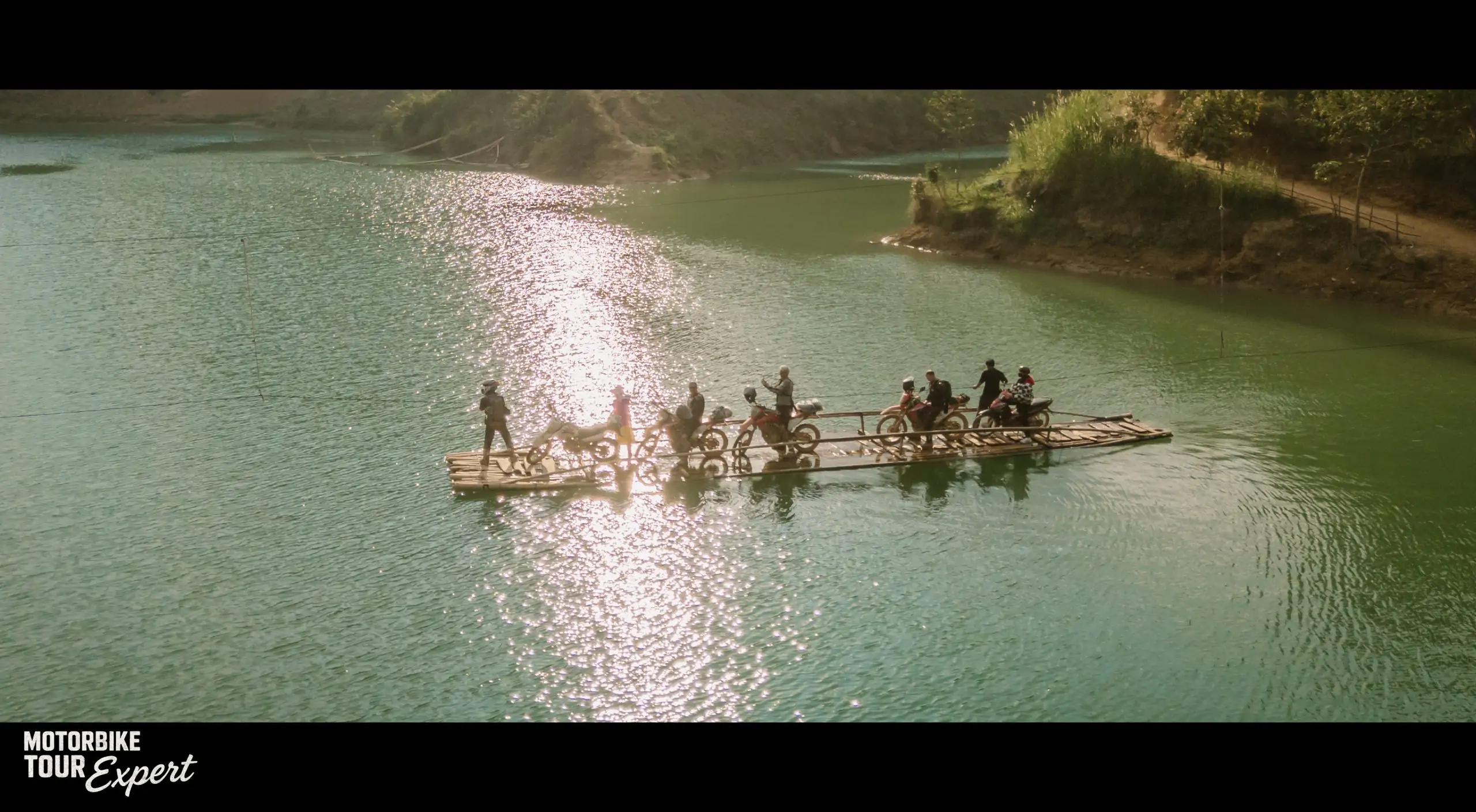 Motorbikes and riders crossing a scenic lake on a wooden raft in Vietnam.
