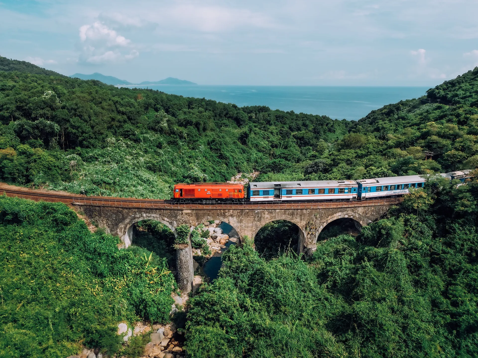 Reunification Express train crossing a stone railway bridge through lush mountains along Vietnam’s coast.
